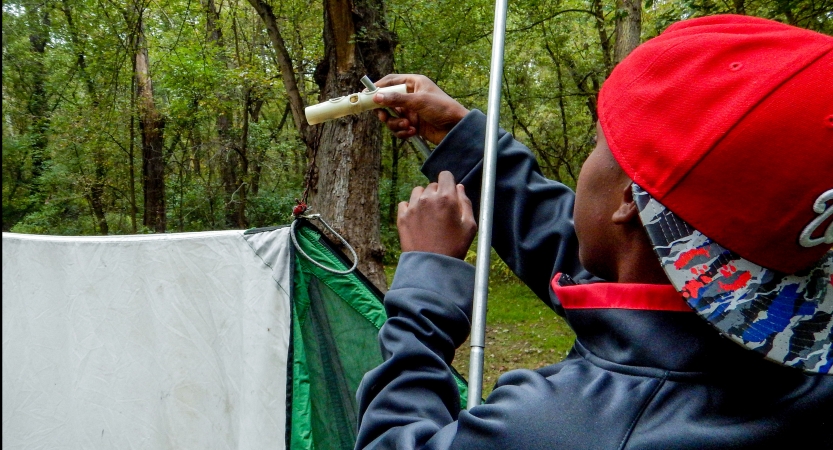 A young person works to tie a knot while assembling a tarp shelter in a wooded area. 